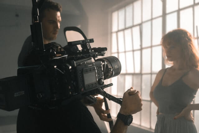 A man holding a large Canon camera and pointing it at a woman stood by a window.