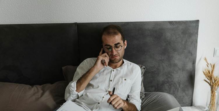 A person sitting on a bed holding a pen and talking on the phone.